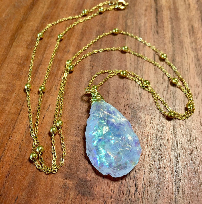 Pu&ograve; includere: Una collana a catena dorata con un grande ciondolo di cristallo iridescente. La catena ha piccole perle dorate.
