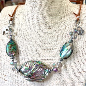 May include: Abalone shell necklace with iridescent colors of green, blue, and purple. The necklace features a large oval abalone pendant and two smaller oval pendants, strung with clear beads and silver-tone metal accents. The necklace is suspended from a brown leather cord.