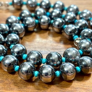 May include: A beaded necklace featuring large, polished, metallic gray spherical beads alternating with small, bright turquoise blue spacer beads. The necklace is arranged in a circular pattern, showcasing the reflective surface of the gray beads.