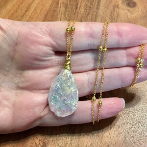 Pu&ograve; includere: Una collana con catena dorata con un grande ciondolo a forma di lacrima in cristallo bianco iridescente.