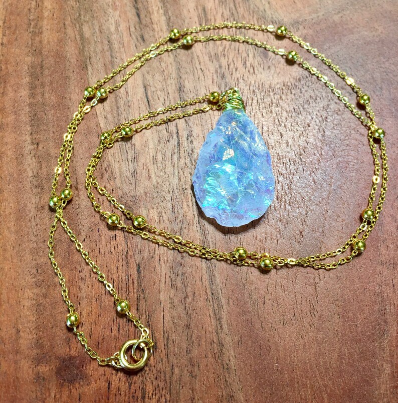 Pu&ograve; includere: Una collana a catena dorata con un ciondolo a forma di lacrima in cristallo blu iridescente. La catena &egrave; decorata con piccole perle dorate.