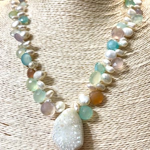 May include: A necklace featuring a variety of colorful gemstones and white pearls. The necklace has a large, irregular-shaped, white, crystal pendant. The gemstones are in shades of blue, green, pink, and orange. The necklace is displayed on a beige surface.