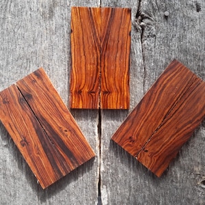 Puede incluir: Tres piezas rectangulares de madera con un rico color marrón rojizo y patrones de vetas intrincados. Las piezas de madera están dispuestas sobre una superficie de madera gris desgastada, mostrando su belleza natural y textura. La madera parece ser de madera dura.