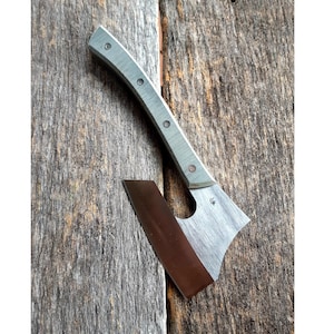 May include: A small, hand-crafted axe with a gray and green handle and a brown and gray blade. The axe is resting on a wooden surface.
