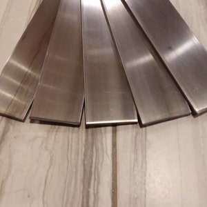 May include: A fan-shaped arrangement of six brushed metal bars. The bars are rectangular, with a flat, reflective surface. The metal has a silver color and a smooth texture. The bars are arranged on a light-colored, patterned surface.