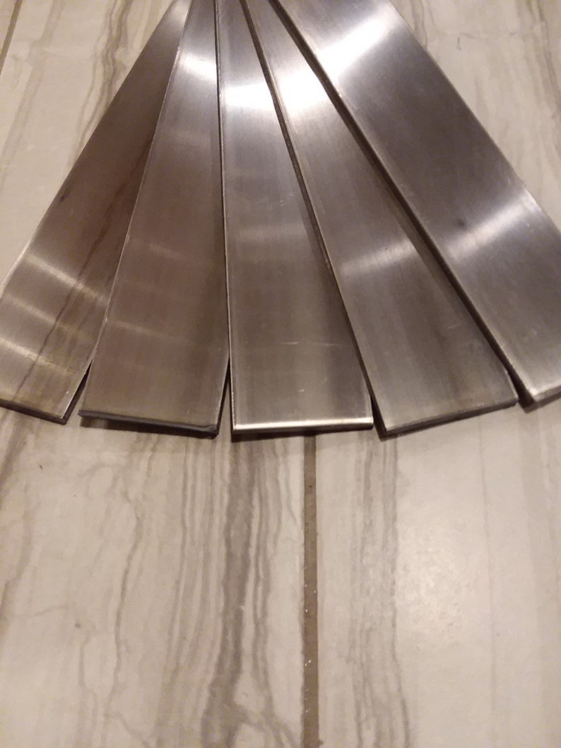 Puede incluir: Disposici&oacute;n en abanico de seis barras de metal cepillado. Las barras son rectangulares, con una superficie plana y reflectante. El metal es de color plateado y tiene una textura lisa. Est&aacute;n sobre una superficie clara y estampada.