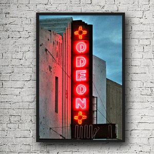 May include: A vintage neon sign with the word "ODEON" in red letters, with a stylized cross design above and below the word. The sign is mounted on a brick building with a faded white paint finish.