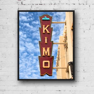 May include: A vintage sign for the Kimo Theatre in Albuquerque, New Mexico. The sign is red with gold lettering and a blue and white eagle design at the top. The sign is against a blue sky with white clouds.