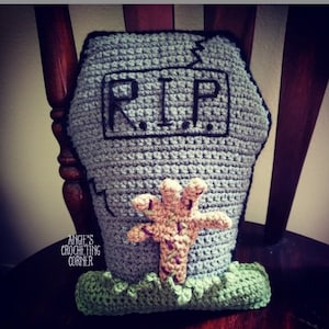 Puede incluir: Una almohada de piedra tombale tejida a crochet con textura de piedra gris, una inscripción "RIP" negra y una base de hierba verde. Una mano de zombie tejida a crochet sobresale del suelo.