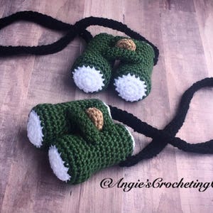 May include: Green crocheted binoculars with a black strap. The binoculars have white accents and a brown detail on the top.