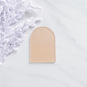 May include: A light pink acrylic blank ornament with a rounded top and a hole for hanging. The ornament is on a white marble surface.