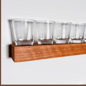 May include: A wooden shot glass holder with six clear shot glasses. The holder is made of reddish-brown wood and is rectangular. The shot glasses are empty and have a simple, straight-sided design. The background is a plain white wall.
