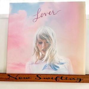 May include: A pink and blue vinyl record with the title "Lover" and a photo of Taylor Swift. The record is displayed on a wooden shelf with the text "Now Swifting" engraved on it.