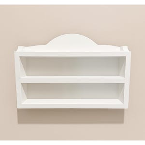 May include: A white, wall-mounted shelf with two shelves and a decorative top. The shelf is rectangular with a curved top and is designed to hold items. The shelf is painted white and is mounted on a beige wall.