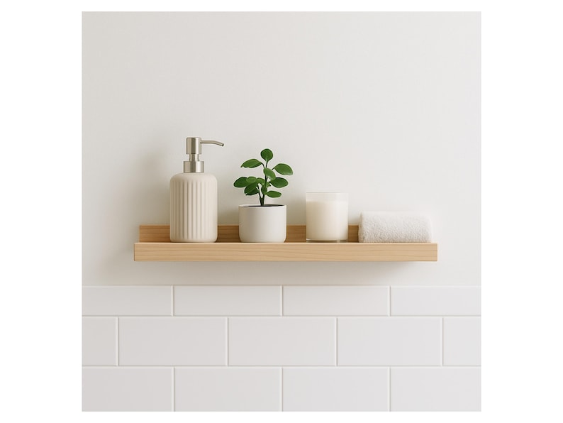 May include: A light wood shelf displays a beige soap dispenser, a small potted plant, a candle, and a folded white towel. The shelf is mounted on a white wall above white subway tiles, creating a minimalist bathroom aesthetic.