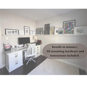 May include: A home office setup with a white desk, chair, and a computer monitor. A shelf above the desk displays framed artwork. The room has a neutral color palette with a rug and dark flooring. Text on the image: Installs in minutes. All mounting hardware and instructions included.