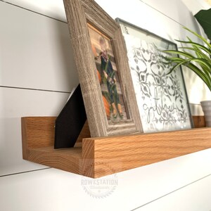 May include: A wooden shelf with a natural finish, featuring two framed pictures and a plant. The shelf is mounted on a white wall. The text "gather" is visible on one of the pictures.