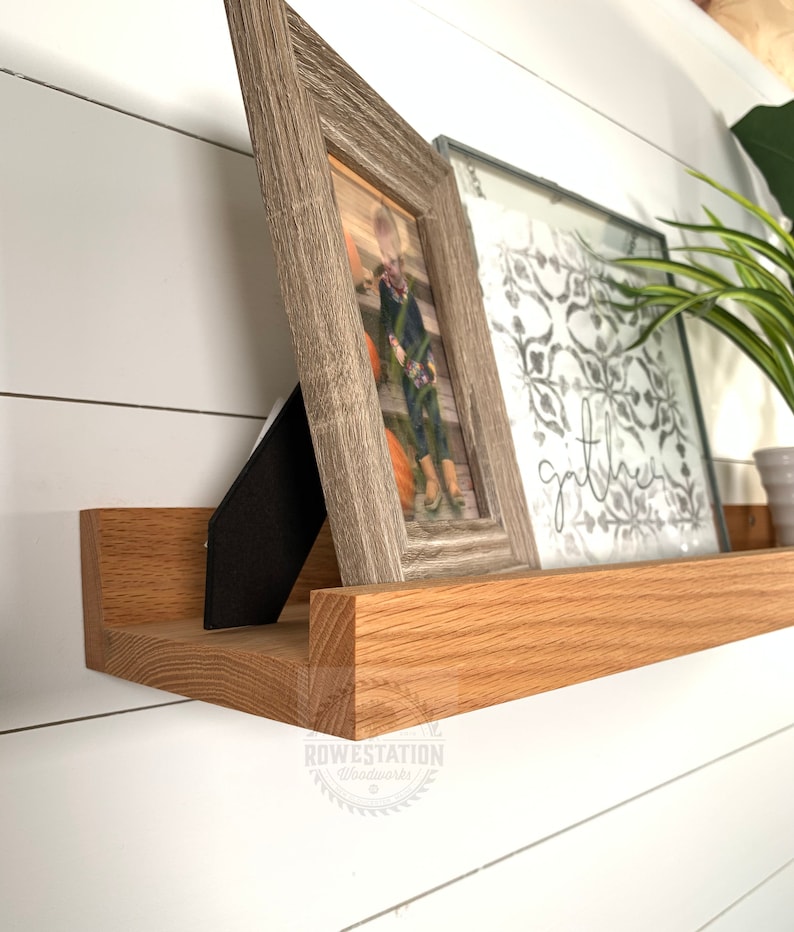 May include: A wooden shelf with a natural finish, featuring two framed pictures and a plant. The shelf is mounted on a white wall. The text "gather" is visible on one of the pictures.