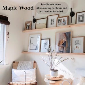 May include: Two wooden shelves with a variety of framed artwork, including a painting of a woman in a yellow dress, and a black and white drawing of a tree. The shelves are mounted on a white wall. The text "Maple Wood" and "Installs in minutes. All mounting hardware and instructions included." is visible in the image.