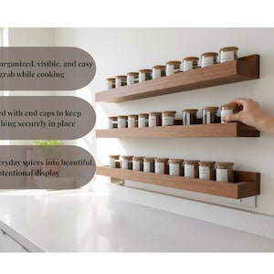 May include: Wooden spice rack with three shelves, each holding several small glass jars with labels and wooden lids. The rack is mounted on a white wall above a countertop. The text on the image says: Keeps jars organized, visible, and easy to grab while cooking.