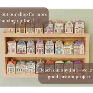 May include: A wooden shelf displays miniature ceramic houses with colorful details. The houses have various roof colors and architectural styles. Text on the image reads, "Check out our shop for more shelving options!" and "Reach out anytime&mdash;we love a good custom project."