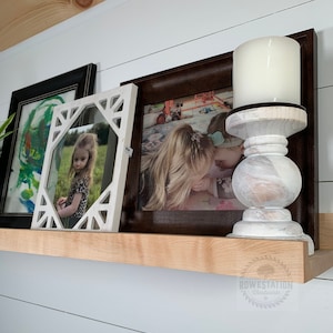 May include: A white wooden shelf with three framed pictures and a white candle holder with a white candle on top. The shelf is mounted on a white wall with wood paneling.