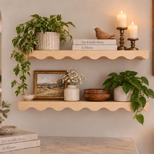 May include: Two light-colored wooden wall shelves with a scalloped front edge. The top shelf holds a potted variegated plant, a small bird figurine, and two stacked books titled The Kinfolk Home At Home, plus two lit candles in brass holders.