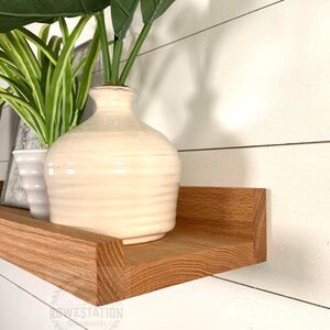 May include: A wooden shelf with a white vase and green plants on top. The shelf is mounted on a white wall with wood paneling. The shelf is angled slightly downward. The text "KOWESTATION Woodworks" is visible in the lower right corner of the image.