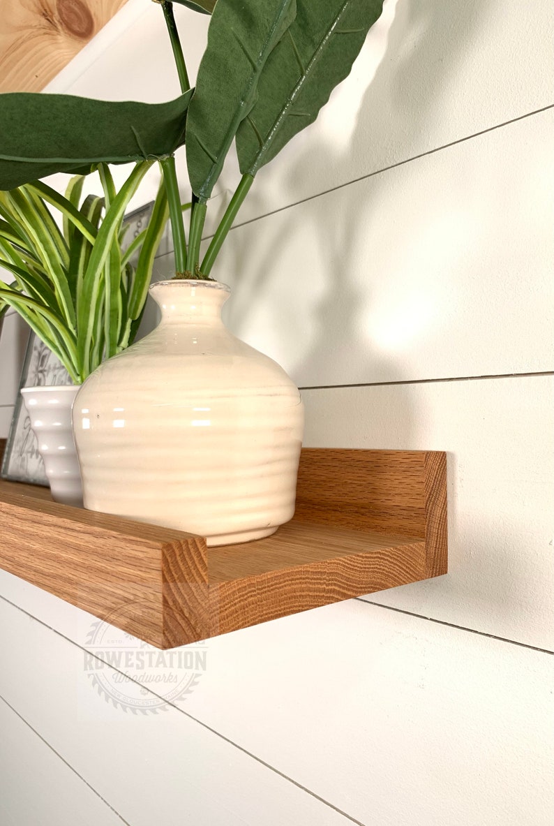 May include: A wooden shelf with a white vase and green plants on top. The shelf is mounted on a white wall with wood paneling. The shelf is angled slightly downward. The text "KOWESTATION Woodworks" is visible in the lower right corner of the image.