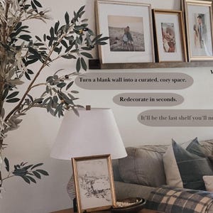 May include: A walnut wood shelf with a white background. The shelf is decorated with framed pictures and a potted olive tree. The text "Turn a blank wall into a curated, cozy space. Redecorate in seconds. It'll be the last shelf you'll need to buy." is written on the wall.