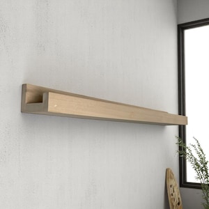 May include: A light-colored, wooden floating shelf mounted on a textured, light gray wall. The shelf has a simple, modern design with a rectangular profile and a small lip. The shelf is empty and ready for display.