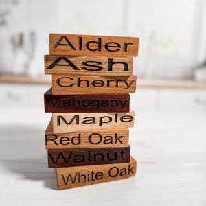 May include: A stack of wooden blocks with different wood types labeled on each block: Alder, Ash, Cherry, Mahogany, Maple, Red Oak, Walnut, White Oak.