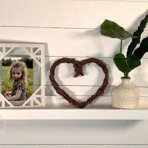 May include: A white shelf displays a framed photo, a heart-shaped wreath, and a cream-colored vase with green leaves. The photo frame has a geometric design. The wreath is made of brown twigs.