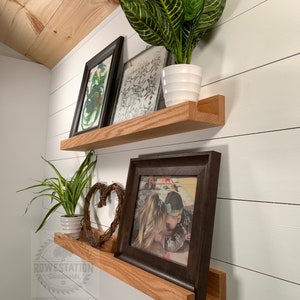 May include: Two wooden shelves with a natural wood finish are mounted on a white wall. The top shelf has a framed print and a potted plant. The bottom shelf has a framed photo, a decorative heart, and a potted plant. The shelves are labeled "ROWESTATION Woodworks".