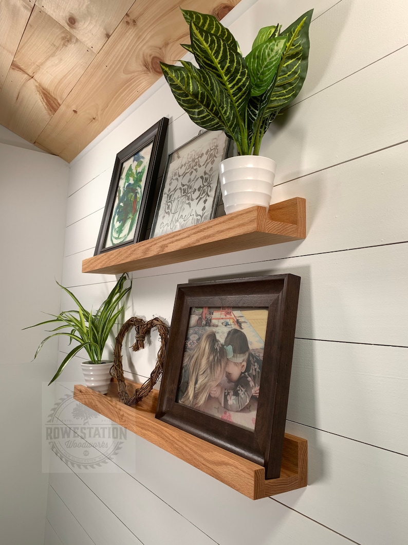 May include: Two wooden shelves with a natural wood finish are mounted on a white wall. The top shelf has a framed print and a potted plant. The bottom shelf has a framed photo, a decorative heart, and a potted plant. The shelves are labeled "ROWESTATION Woodworks".