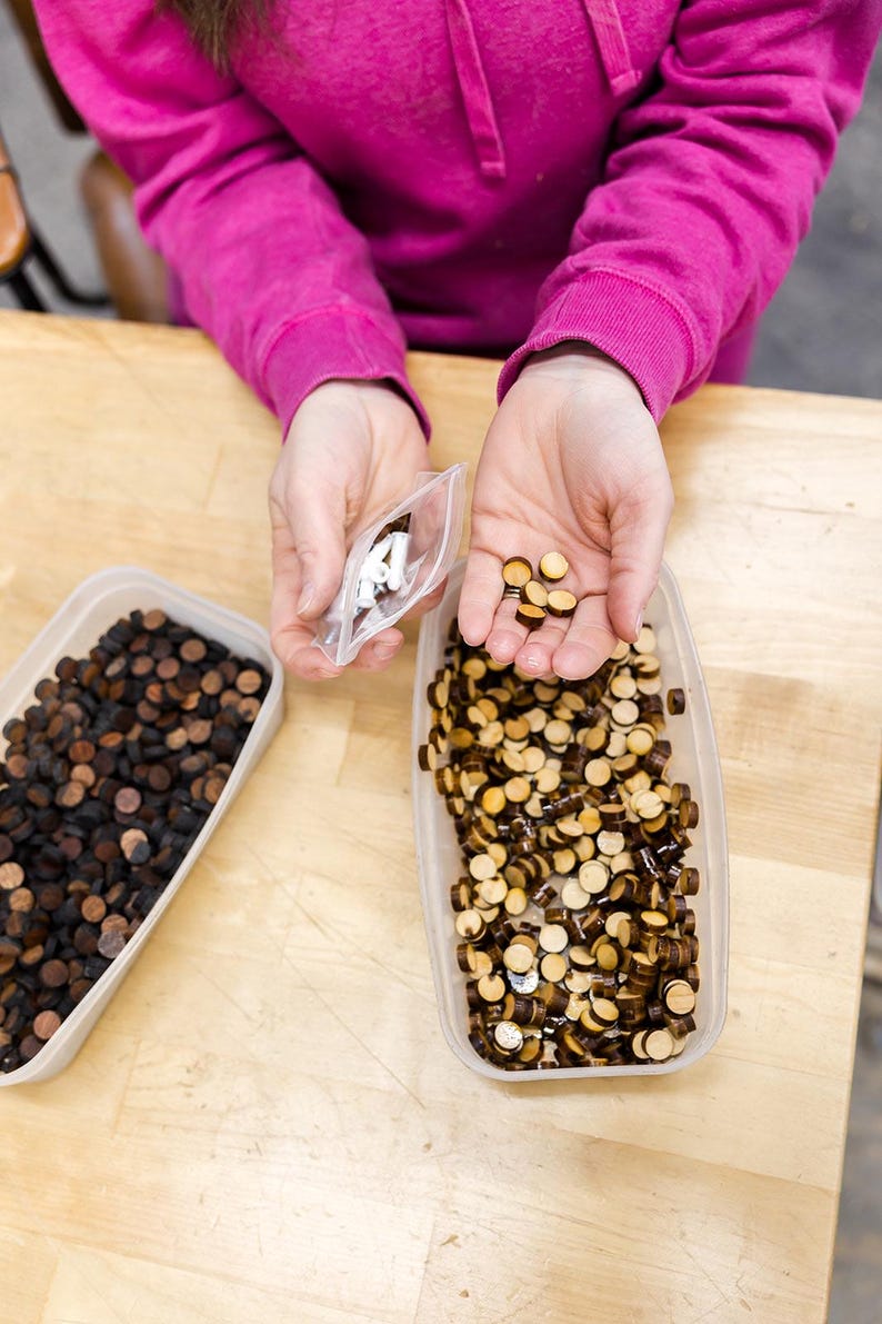 May include: Two containers filled with small wooden circles, one container is filled with dark brown circles and the other with light brown and dark brown circles. A person is holding a small bag of white circles.
