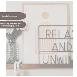 May include: A framed artwork with a neutral color palette, featuring a geometric design and the words "RELAX AND UNWIND" in black metal lettering. A vase with flowers and decorative beads are on a wooden shelf below.