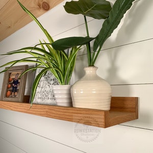May include: A wooden shelf with a rounded edge, mounted on a white wall. The shelf is decorated with a framed photo, a potted plant, and a vase with a leafy plant. The shelf is labeled "Rowestation Woodworks".