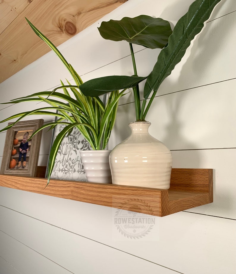 May include: A wooden shelf with a rounded edge, mounted on a white wall. The shelf is decorated with a framed photo, a potted plant, and a vase with a leafy plant. The shelf is labeled "Rowestation Woodworks".