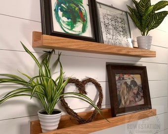 May include: Two wooden shelves with a white background. The top shelf has a black framed painting, a white framed print with the word "gather" on it, and a potted green plant. The bottom shelf has a potted green plant, a brown framed photo, and a woven heart.