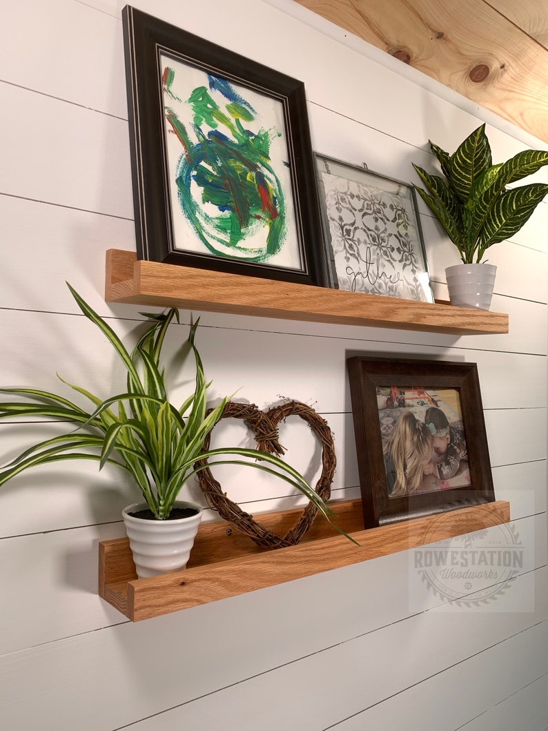 May include: Two wooden shelves with a white background. The top shelf has a black framed painting, a white framed print with the word "gather" on it, and a potted green plant. The bottom shelf has a potted green plant, a brown framed photo, and a woven heart.