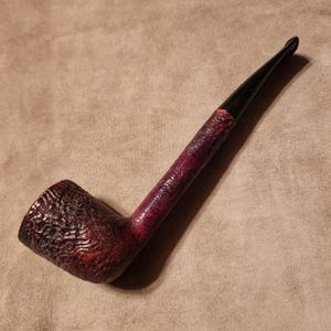 Pode incluir: Um cachimbo de tabaco com uma piteira escura e texturizada e um canudo que transita de um bordô escuro para preto. O cachimbo tem um formato clássico e está sobre um fundo neutro.