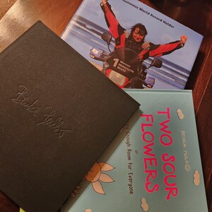 May include: Three books stacked on top of each other. The top book is a blue and white book with the text "Guinness World Record Holder" and a photo of a woman on a motorcycle. The middle book is black with the text "Bela Pulks" in white. The bottom book is blue with the text "Two Sour Flowers" in pink and white.
