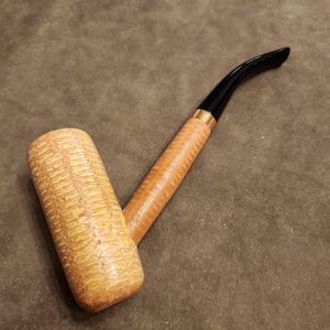May include: A corn cob pipe with a light brown bowl and a curved black stem. The stem is connected to the bowl with a light brown wooden shank and a gold-colored band. The bowl has a textured surface.