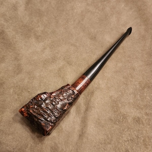 May include: A handcrafted tobacco pipe with a dark brown, textured bowl and a long, black stem. The pipe's bowl has a rough, carved appearance, and the stem is smooth and glossy. The pipe is designed for smoking tobacco.