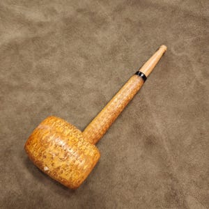 May include: A tobacco pipe crafted from light brown, textured wood with a cylindrical bowl and a long, slender stem. The stem features a smooth, tapered mouthpiece separated from the main body by a thin, distinct black band near the center.