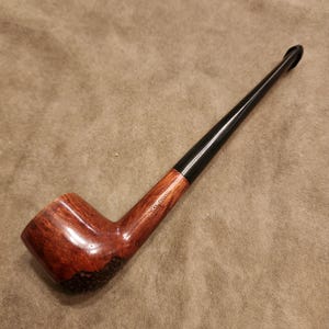 Peut inclure: Une pipe à tabac classique avec un foyer brun foncé et un long tuyau noir. La pipe a un design incurvé et une finition lisse et polie. Le foyer est brun clair avec une base texturée plus foncée. La tige est reliée au foyer par une pièce en bois.