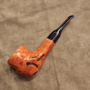 May include: A wooden tobacco pipe with a curved stem and a dark, glossy mouthpiece. The bowl has a light brown finish with dark etched details. The pipe is designed for smoking tobacco.