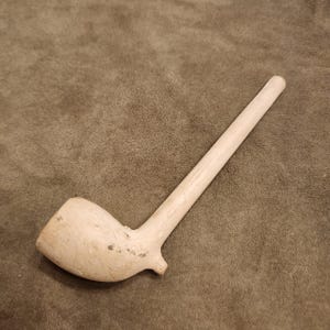 Puede incluir: Una pipa de fumar de madera sin terminar en color beige claro, con un hornillo redondeado y liso y un tallo largo y recto. La superficie de la pipa muestra patrones de veta de madera natural y variaciones sutiles de color, con un diseño simple.