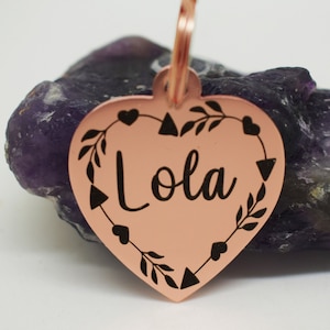 Puede incluir: Una etiqueta para mascotas en forma de corazón de cobre con un diseño grabado en negro que presenta hojas, corazones y el nombre "Lola".